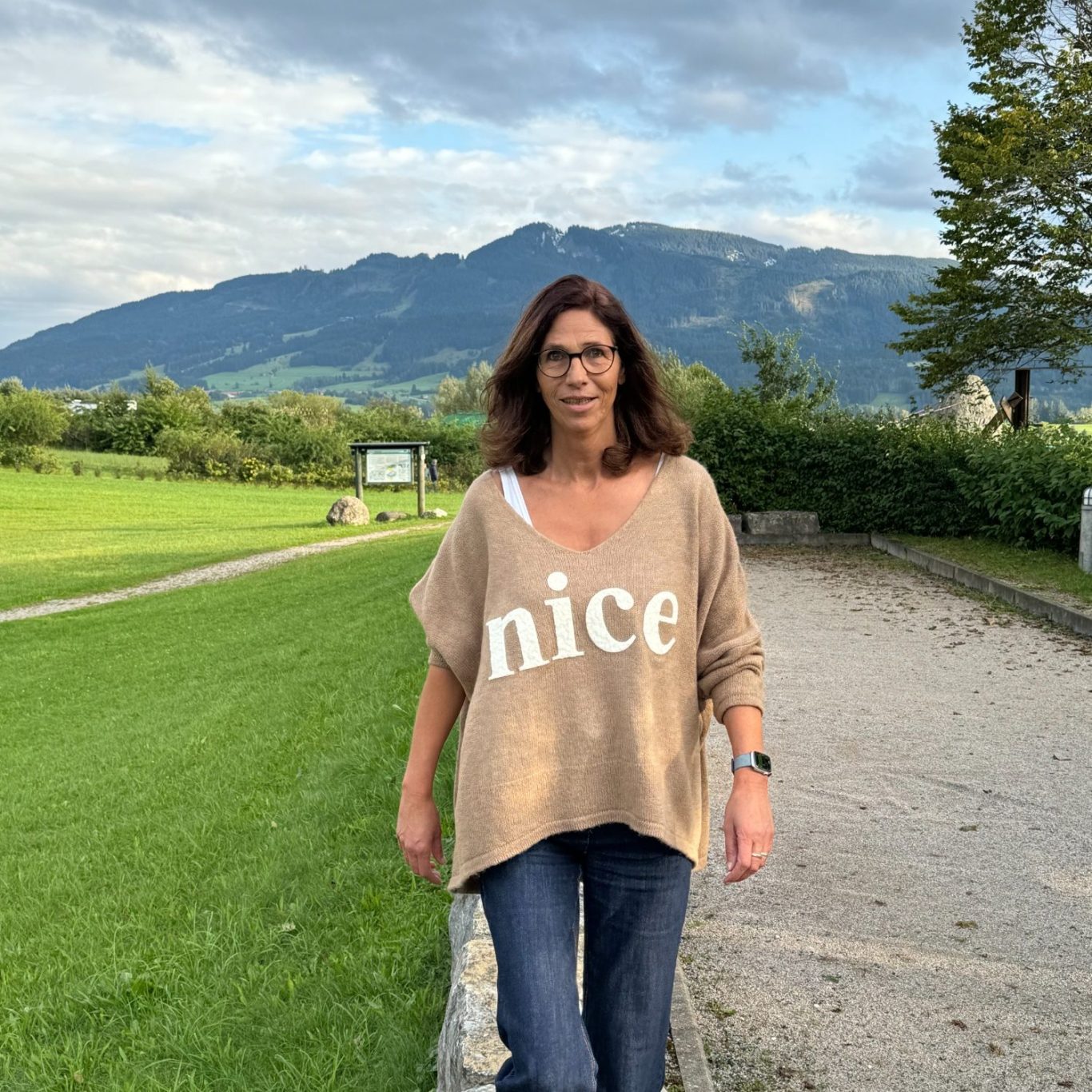 Heike Votteler steht lächelnd und balanciert auf einer niedrigen Steinmauer in einer Berglandschaft. Sie trägt eine beigen Pullover 