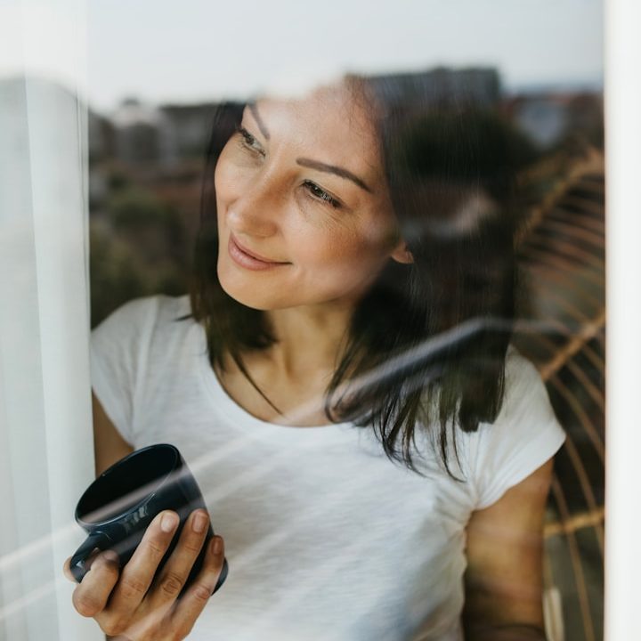 Frau mit weißem Shirt am Fenster und einer Tasse in der Hand